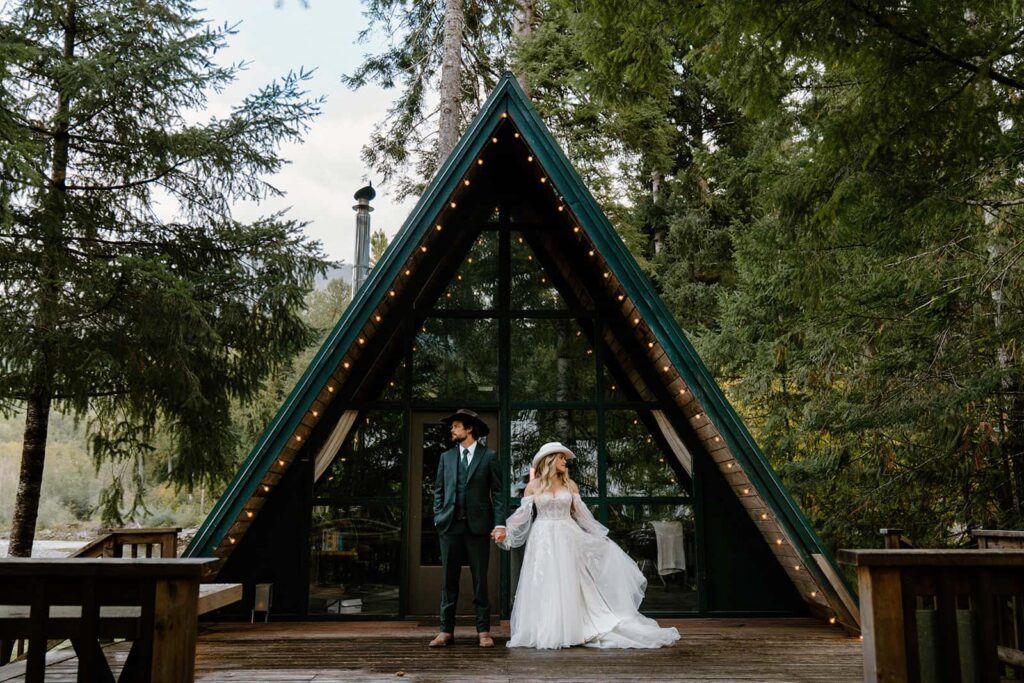 Groom in dark suit with cowboy boots and hat holding bride's hand in a lacey wedding dress and cowgirl hat in from of an a-frame cabin in Mount Rainier.