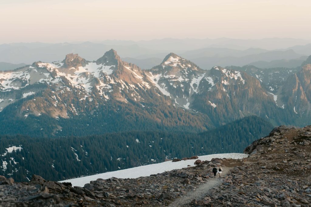 Bride and groom in wedding attire and hiking packs in the distance while on a trail at Mount Rainier National Park. There is snow and part of the Cascades in the background. 