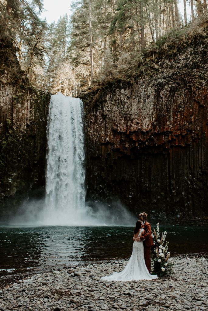 Bride and groom kissing at their ceremony location next to a boho floral standing piece and in front of a large waterfall in Oregon state.