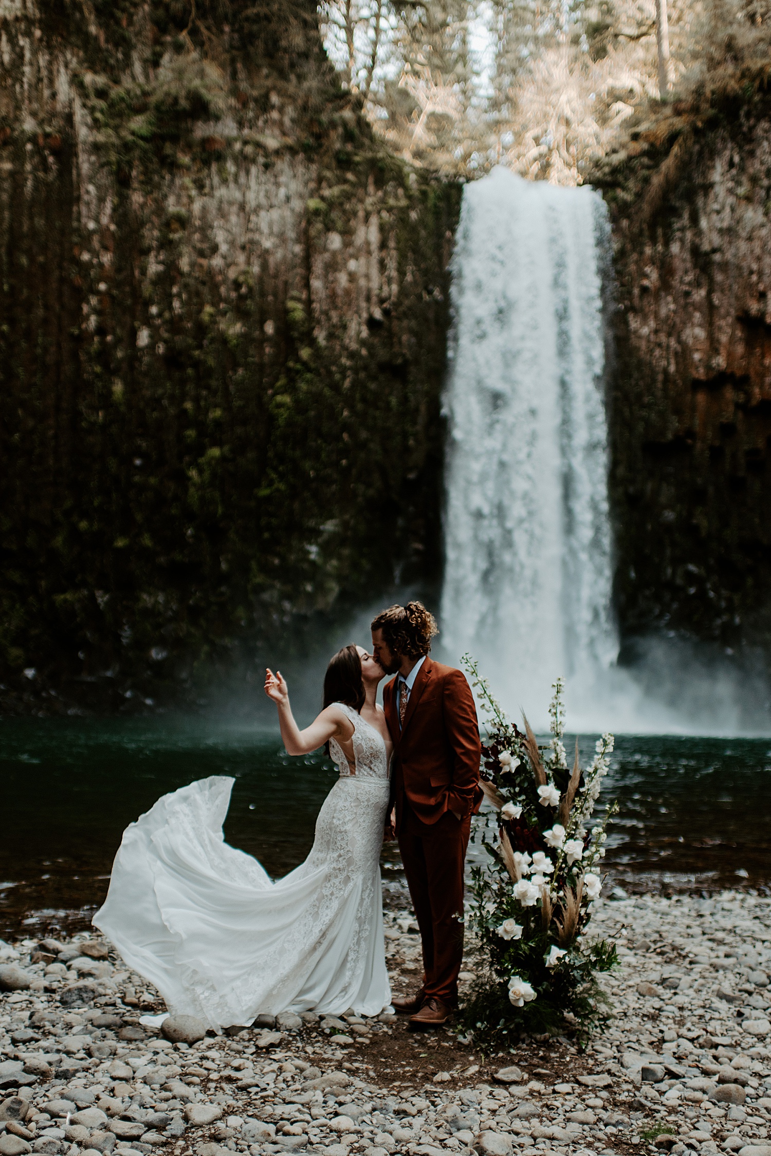 Groom kissing his bride in front of a waterfall while she flicks her boho wedding train in the air.