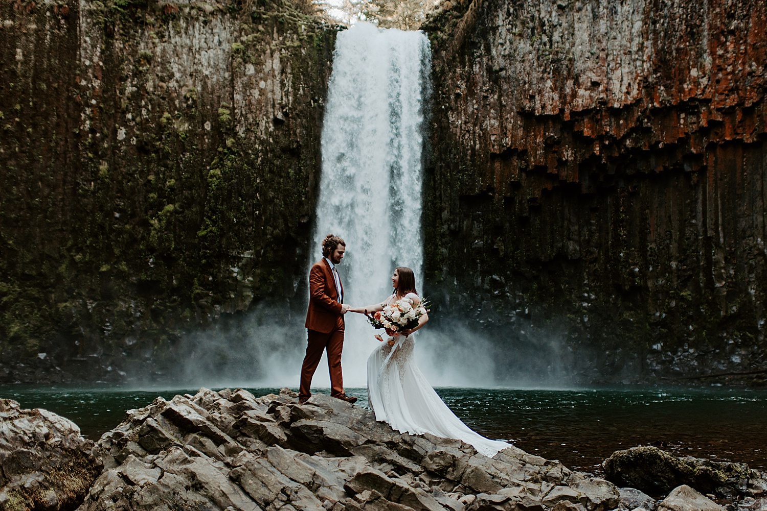 Bride and groom posing on large rocks in front of an Oregon waterfall wearing a lacey wedding dress and a burnt orange suit, and holding a boho bridal bouquet.