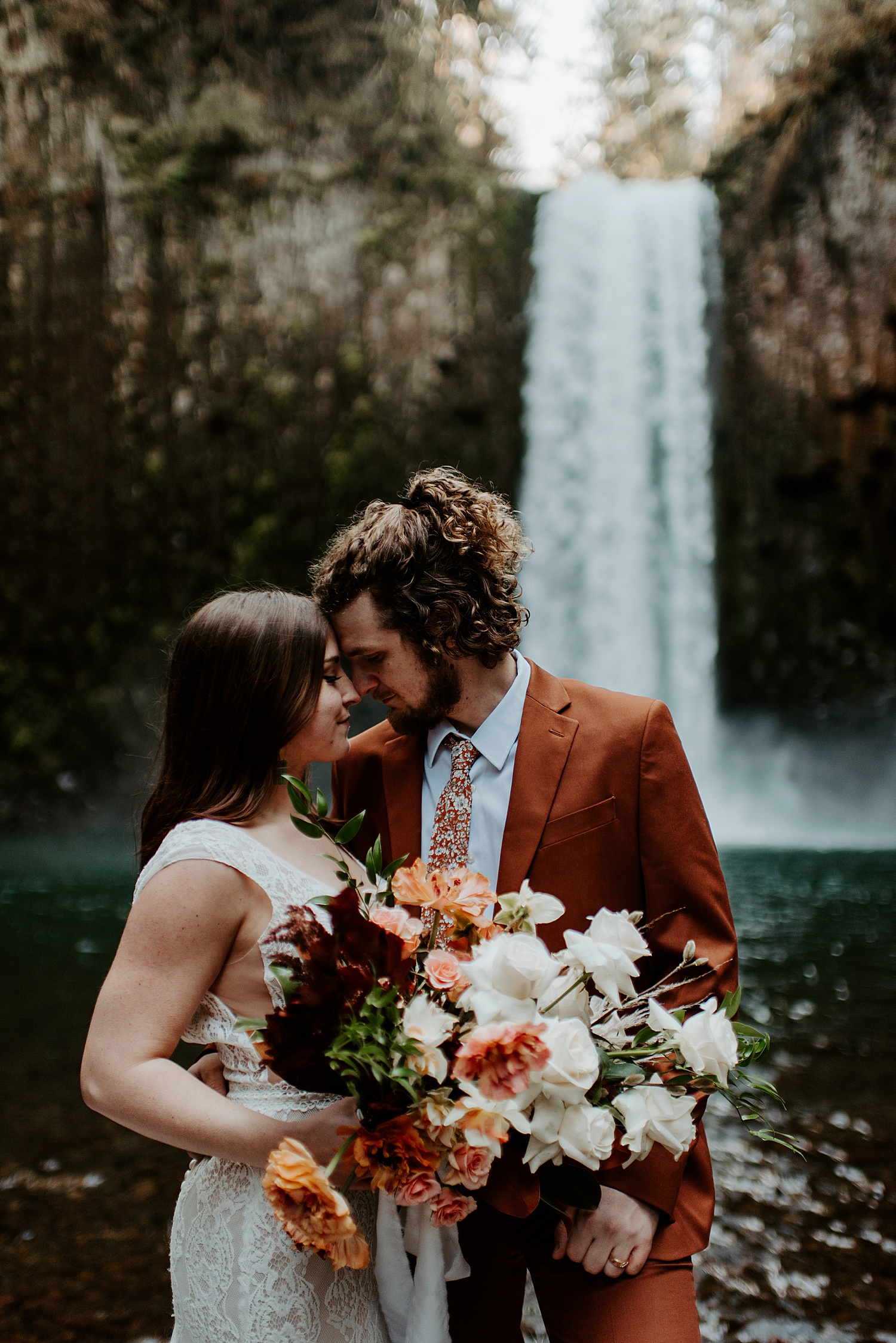 Newly married couple resting their heads together and holding a boho bridal bouquet in front of a waterfall in Oregon.