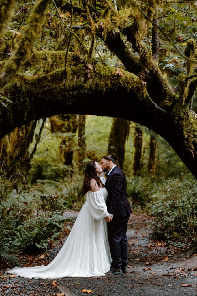 Bride and groom in wedding attire kissing under a curved tree in the Hoh Rainforest of Olympic National Park, Washington.