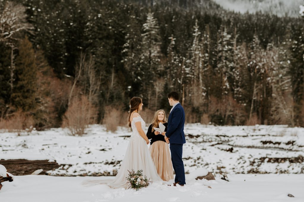 Bride and groom saying their vows with an officiant in the winter on a snowy riverbank.
