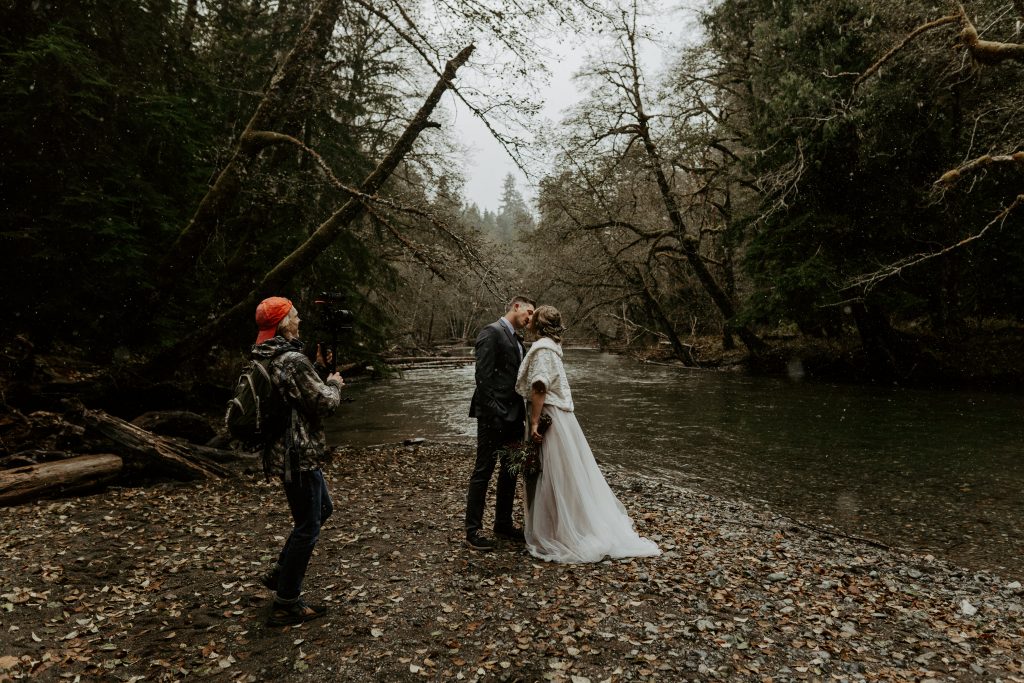 Married couple kissing by the river in the snow with Adam, an elopement videographer, capturing video. 