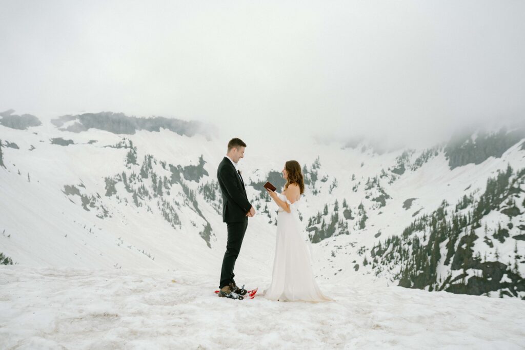 Bride and groom in wedding attire and snow shoes saying vows on a snowy mountain with a cloudy background in the North Cascades. 