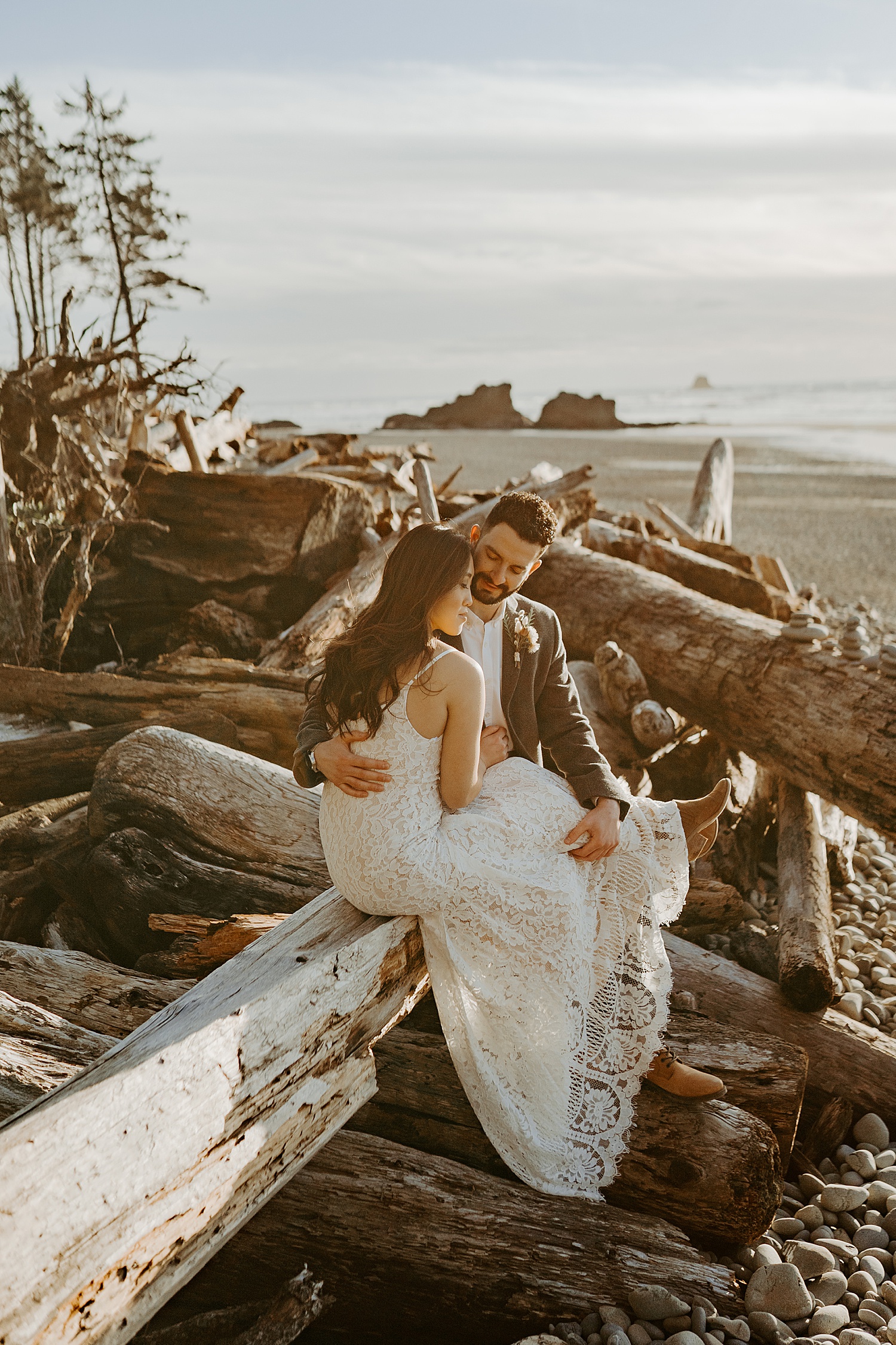 married couple sitting in their wedding attire on logs at the beach