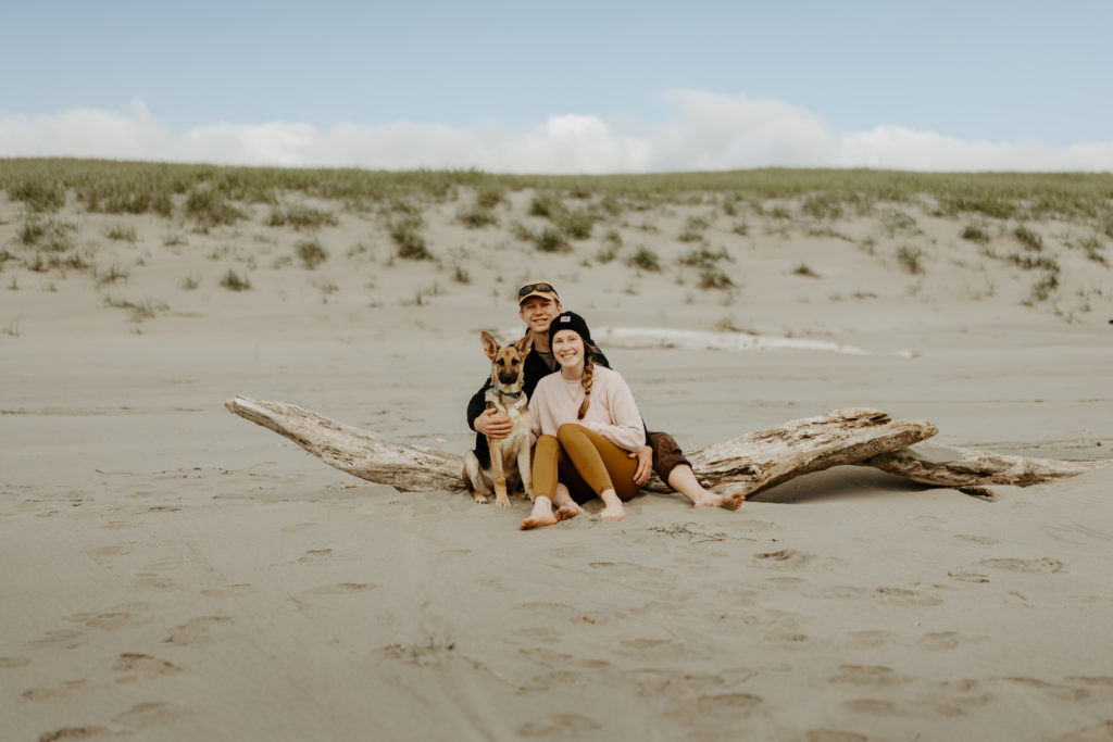 Male and female couple sitting on a piece of drift wood in the sand with their German Shepherd puppy. 