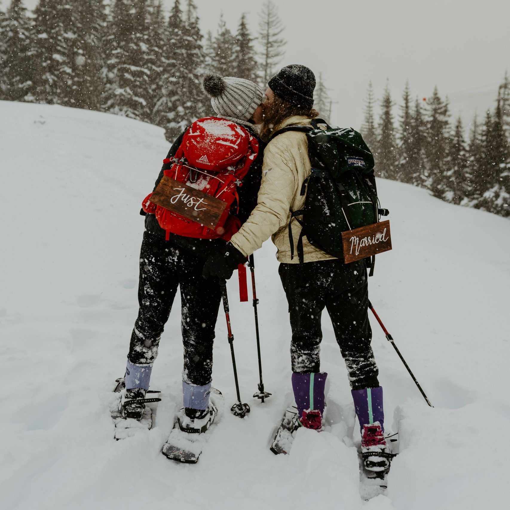 Couple kissing in the snow while snowshoeing with "Just Married" signs on backpacks