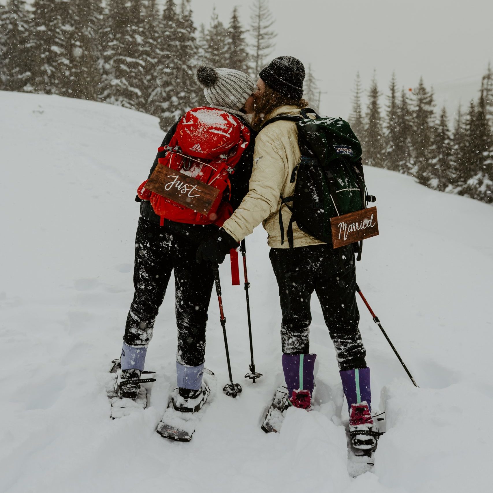 Couple kissing in the snow while snowshoeing with "Just Married" signs on backpacks