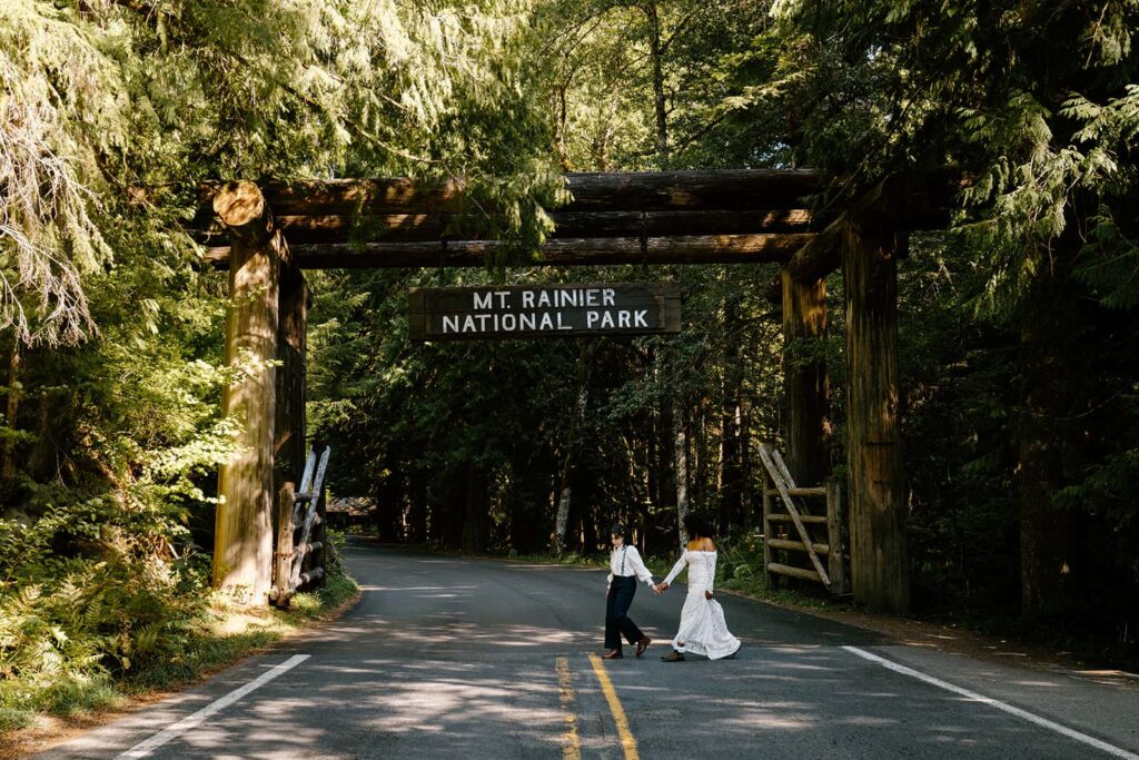Two brides in wedding attire holding hands while walking in the street in front of the Nisqually Entrance to Mount Rainier National Park. 