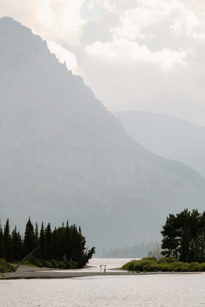 Far away photo of a couple twirling on a part of the beach surrounded by water with trees and a massive mountain in the background at Pray Lake, Montana
