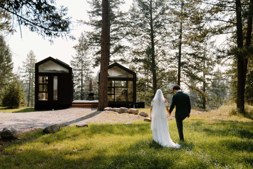 Bride and groom walking in the grass towards two small black tiny homes in the forest. 