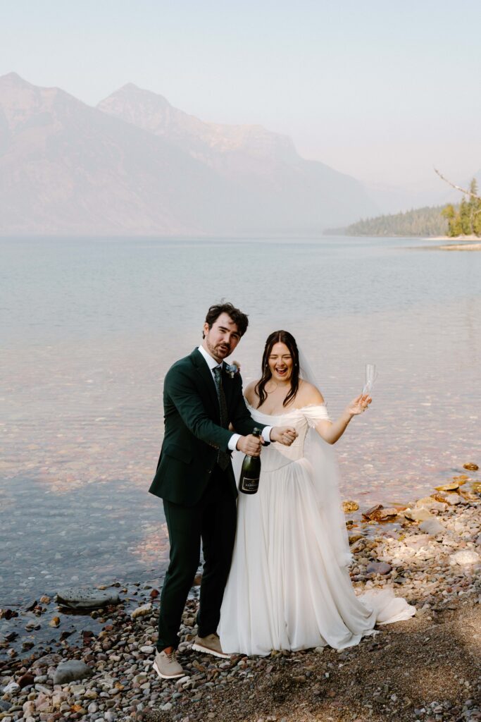 Photo of a bride and groom in wedding attire popping champagne on the shore of Lake McDonald after their Glacier National Park elopement ceremony. 