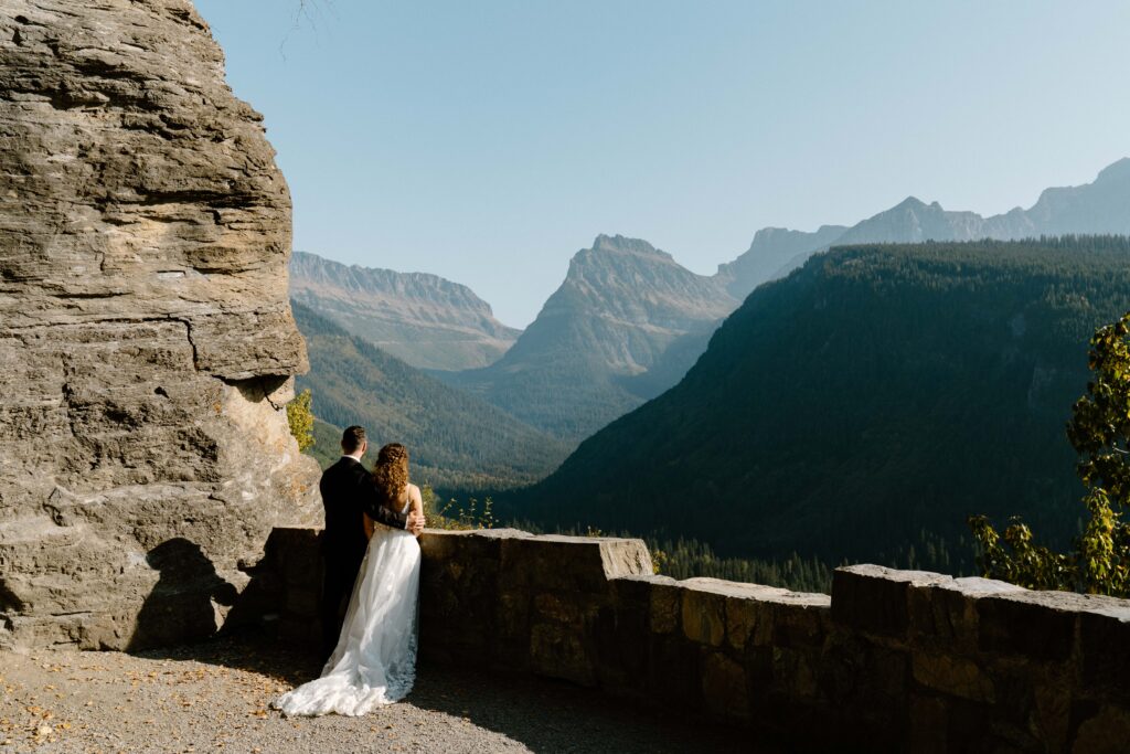 Bride and groom in wedding attire at an overlook along the Going-to-the-Sun Road in Glacier National Park facing away from the camera and looking at the mountains. 