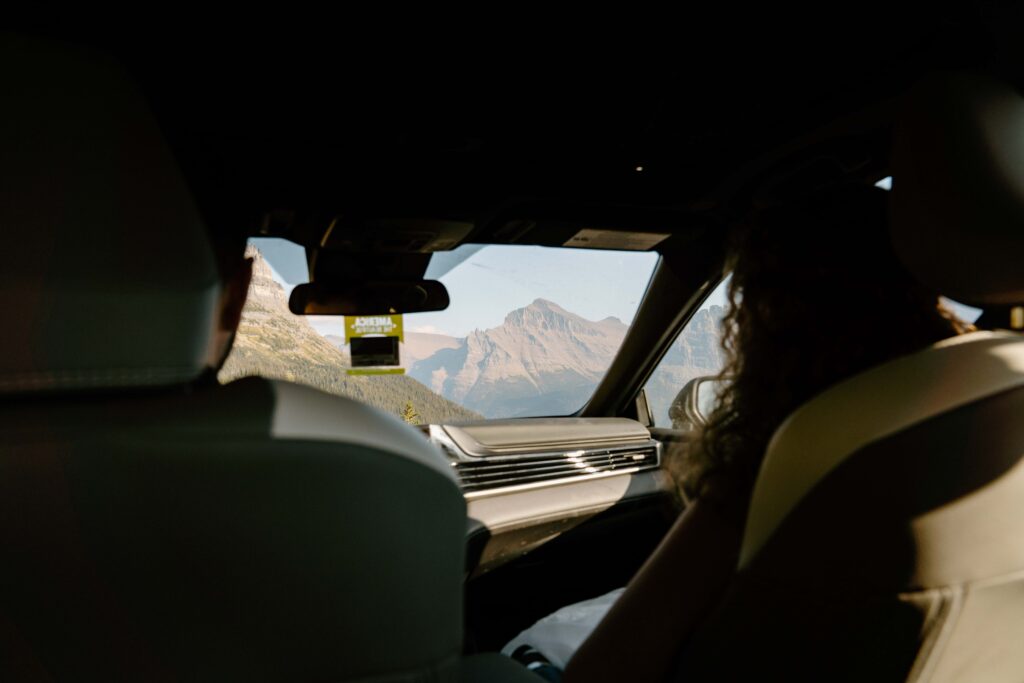 Image from the backseat of a car with the couple in front driving and views of big mountains through the windshield 