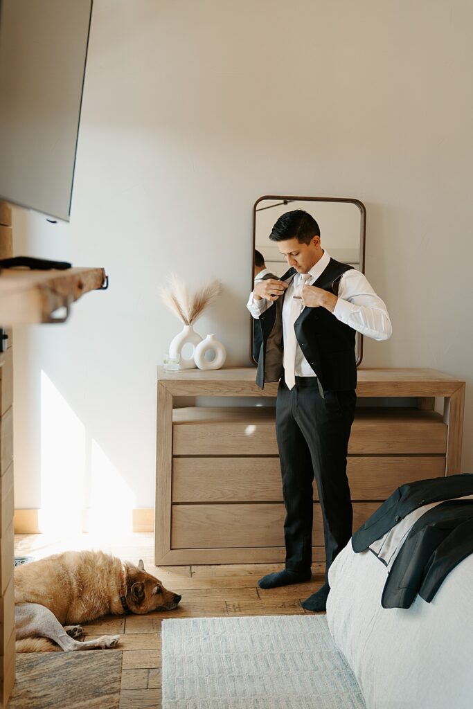 Groom putting on a vest and tie in a bedroom with his dog laying on the floor in front of him. 