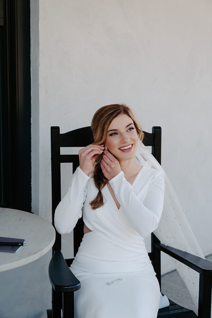 Beautiful bride in a sleek, long sleeve wedding dress sitting down and putting her earrings on. 