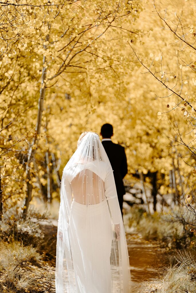 Image of a bride with a pearly veil walking towards the groom with both of their backs turned to the camera. They are sharing a first look on their wedding day under the yellow aspen trees. 