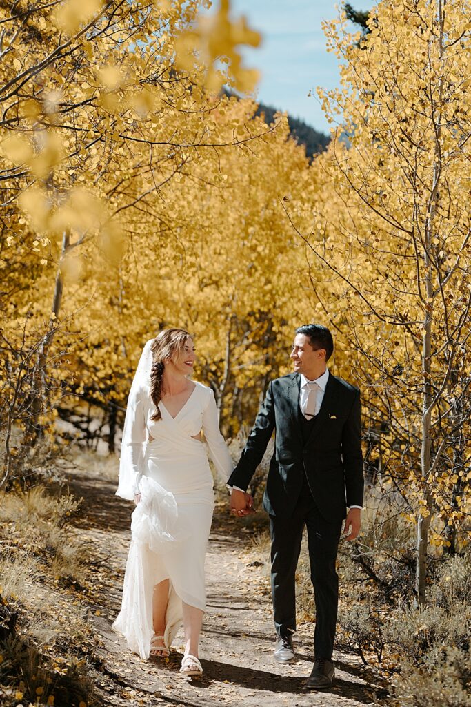 Bride and groom in wedding attire, holding hands while walking under the yellow aspen trees. 