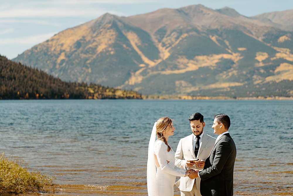 Bride and groom holding hands during their wedding ceremony with Twin Lakes in the background and fall colors in the Colorado mountains. 