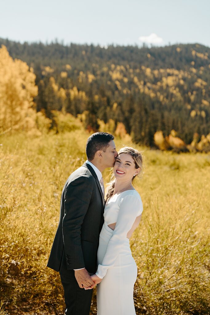 Groom kissing bride on the cheek with yellow fall colors behind them. 