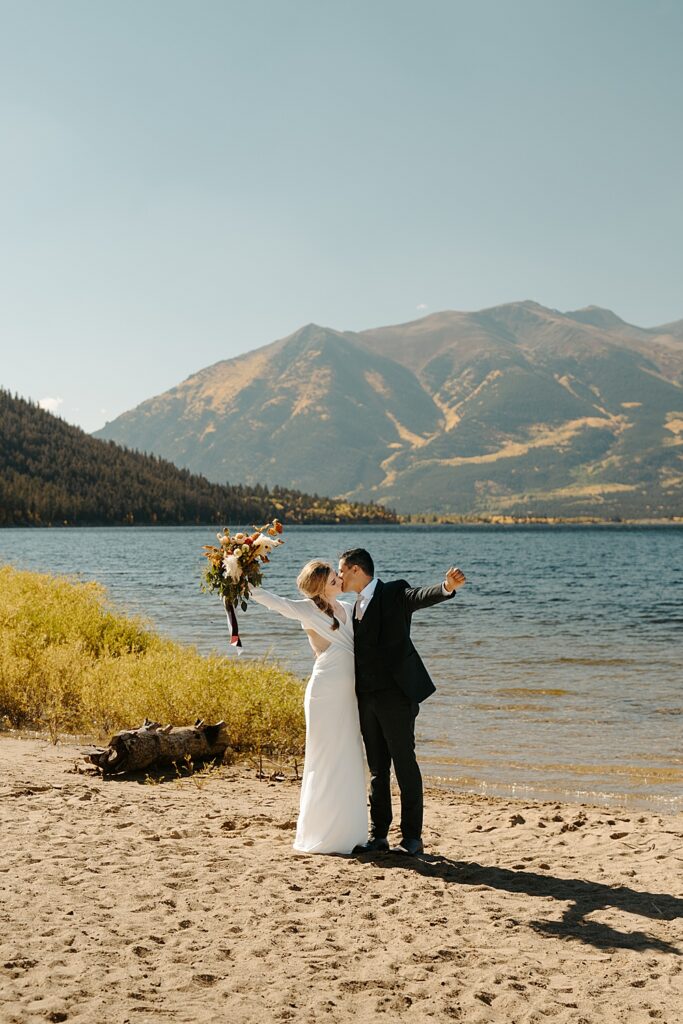 Bride and groom kissing while cheering with their arms in the air. The background is Twin Lakes, Colorado with yellow fall colors on the mountain. 