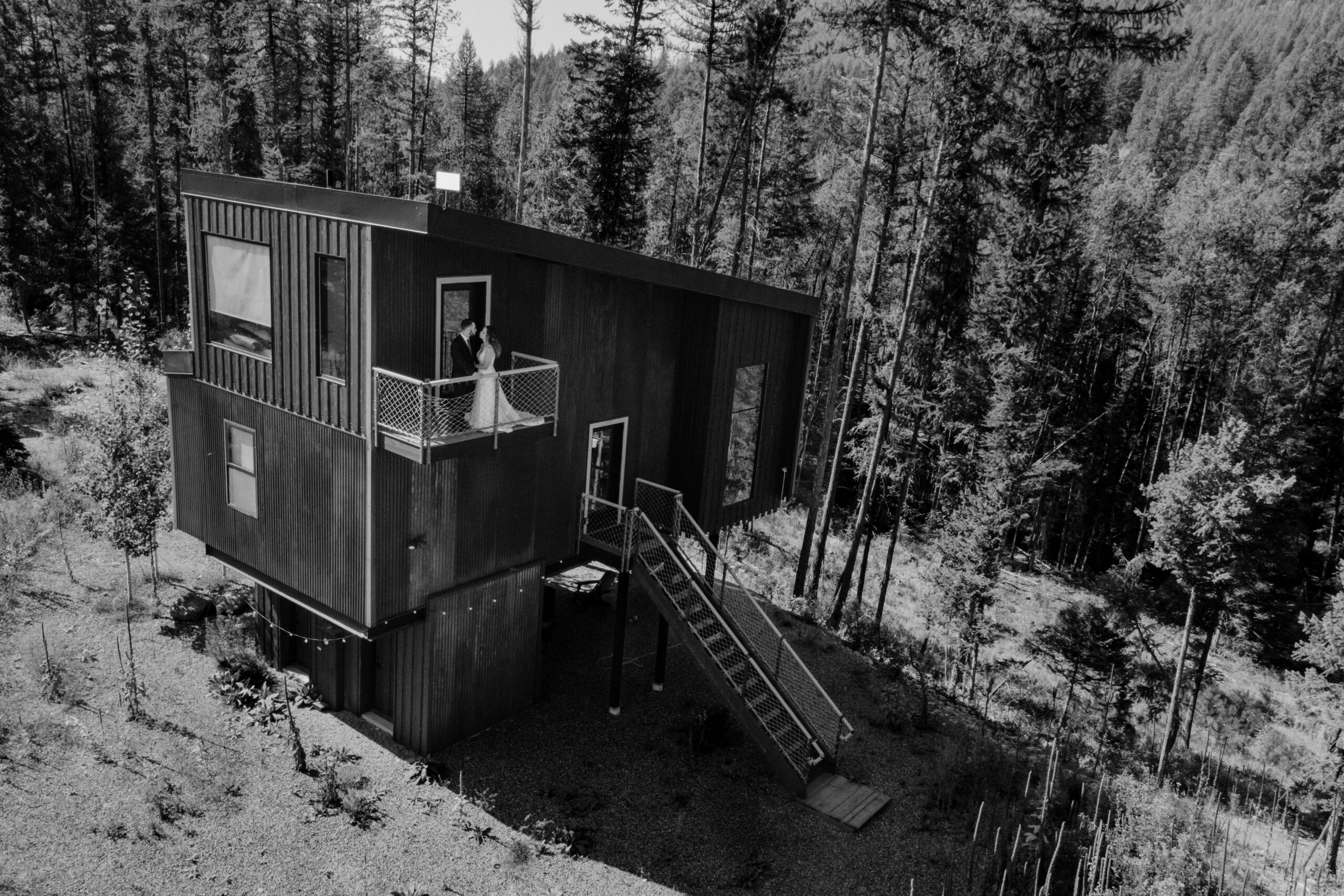 Black and white drone photo of a bride and groom standing on the balcony of a unique rental cabin in the woods. 