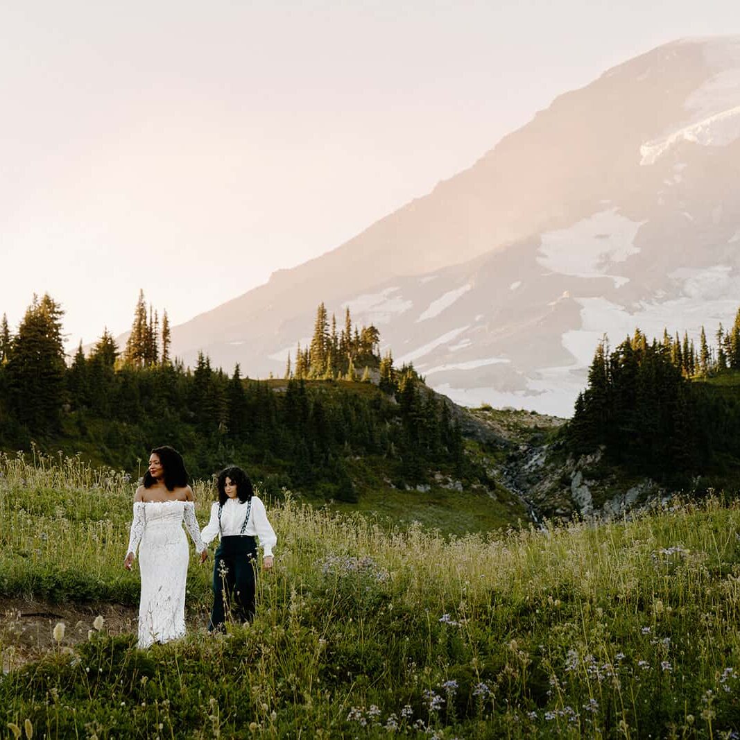 Two brides in wedding attire holding hands walking down a trail in the summer at Mount Rainier National Park. 