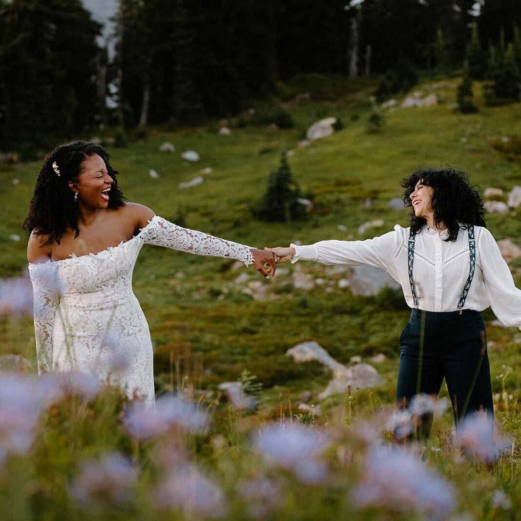 Up close image of two brides in wedding attire holding hands and smiling with purple wildflowers in the foreground. 