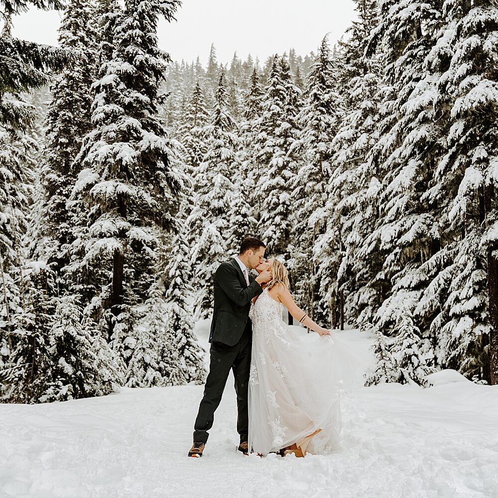 Bride and groom in wedding attire kissing in a winter wonderland with snow and trees in Washington State. 