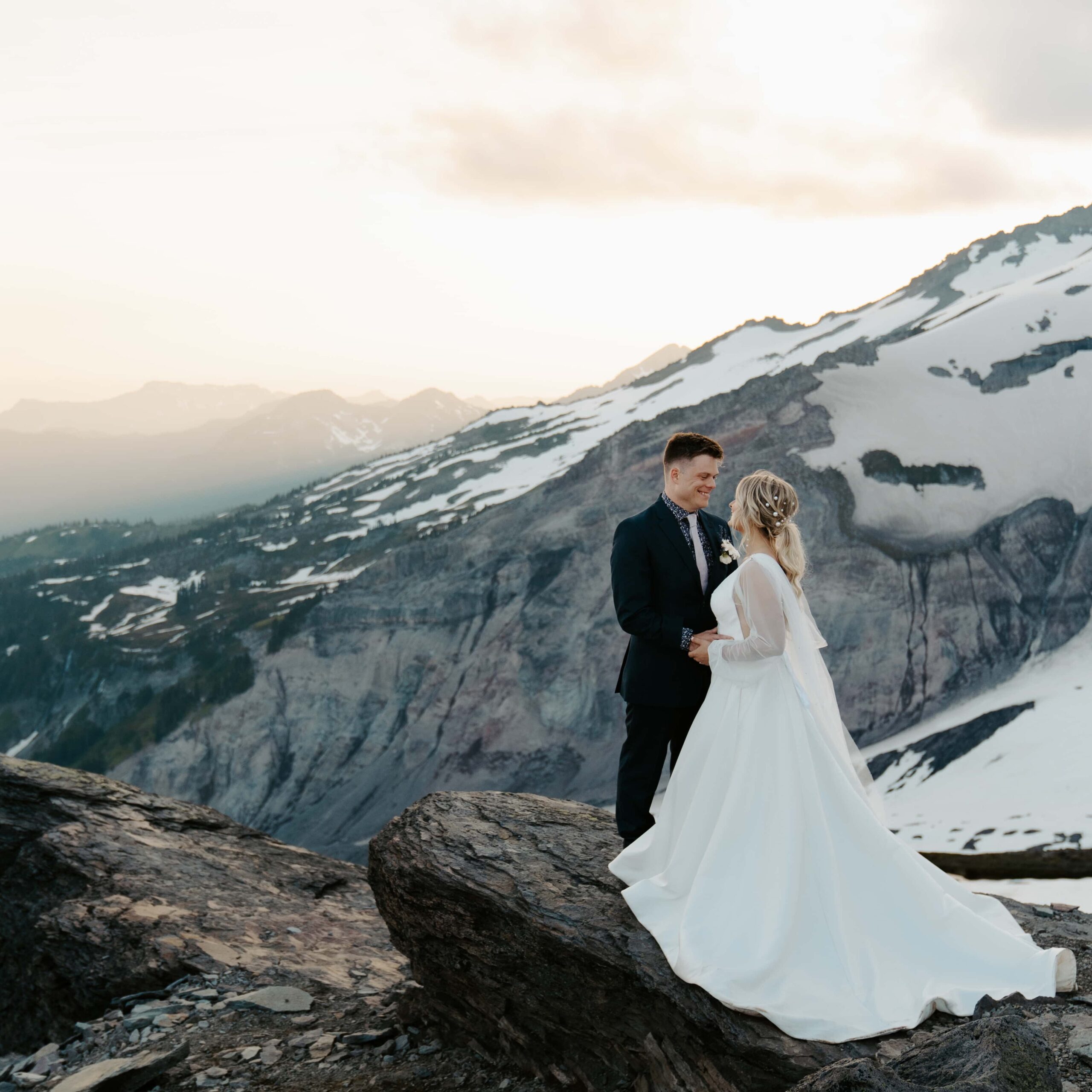 Bride and groom in wedding attire standing on a rock after sunset with Mount Rainier in the background. 