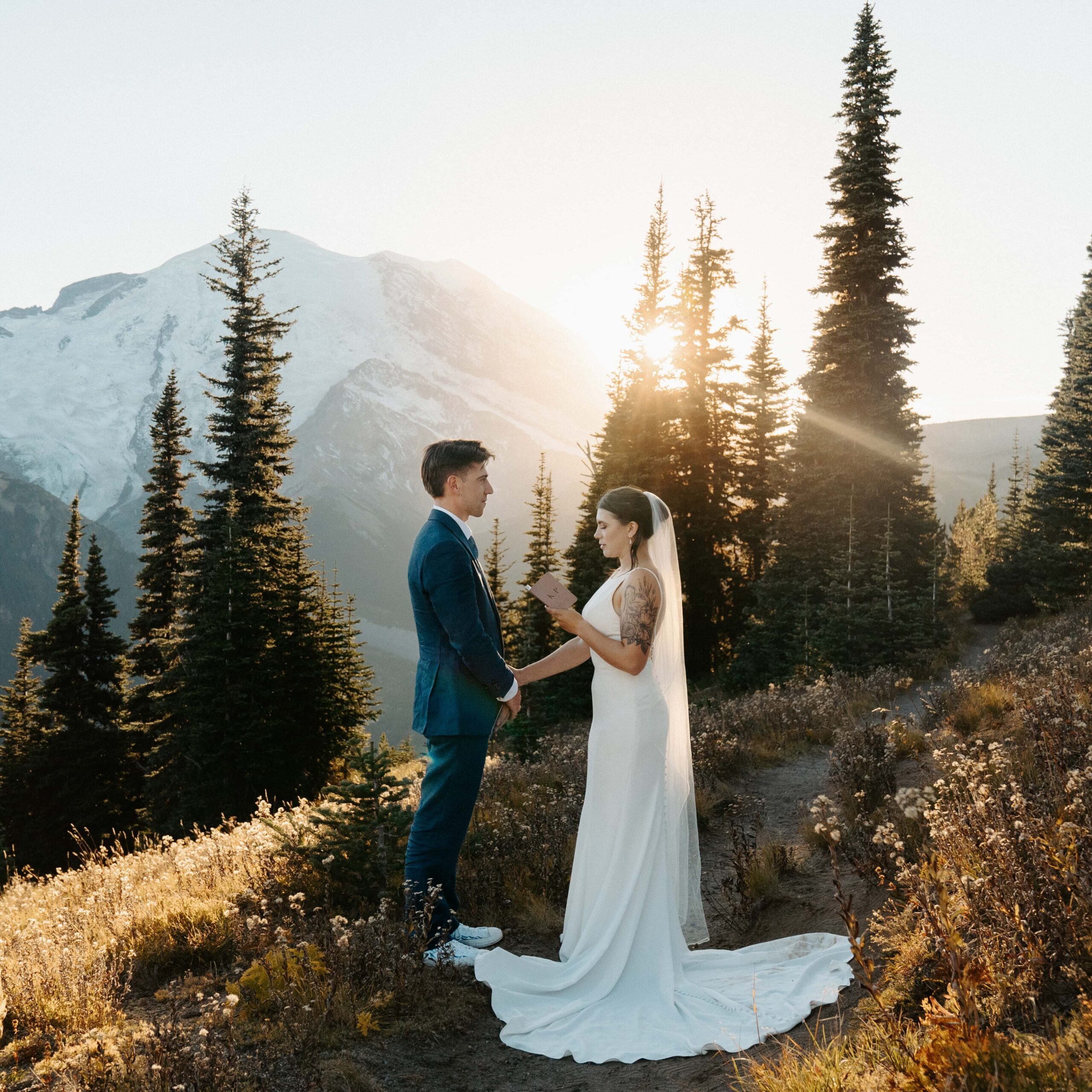 Bride and groom in wedding attire standing on a trail at sunset, saying their vows with Mount Rainier in the background. The image was taken in the fall with fall foliage in the park. 