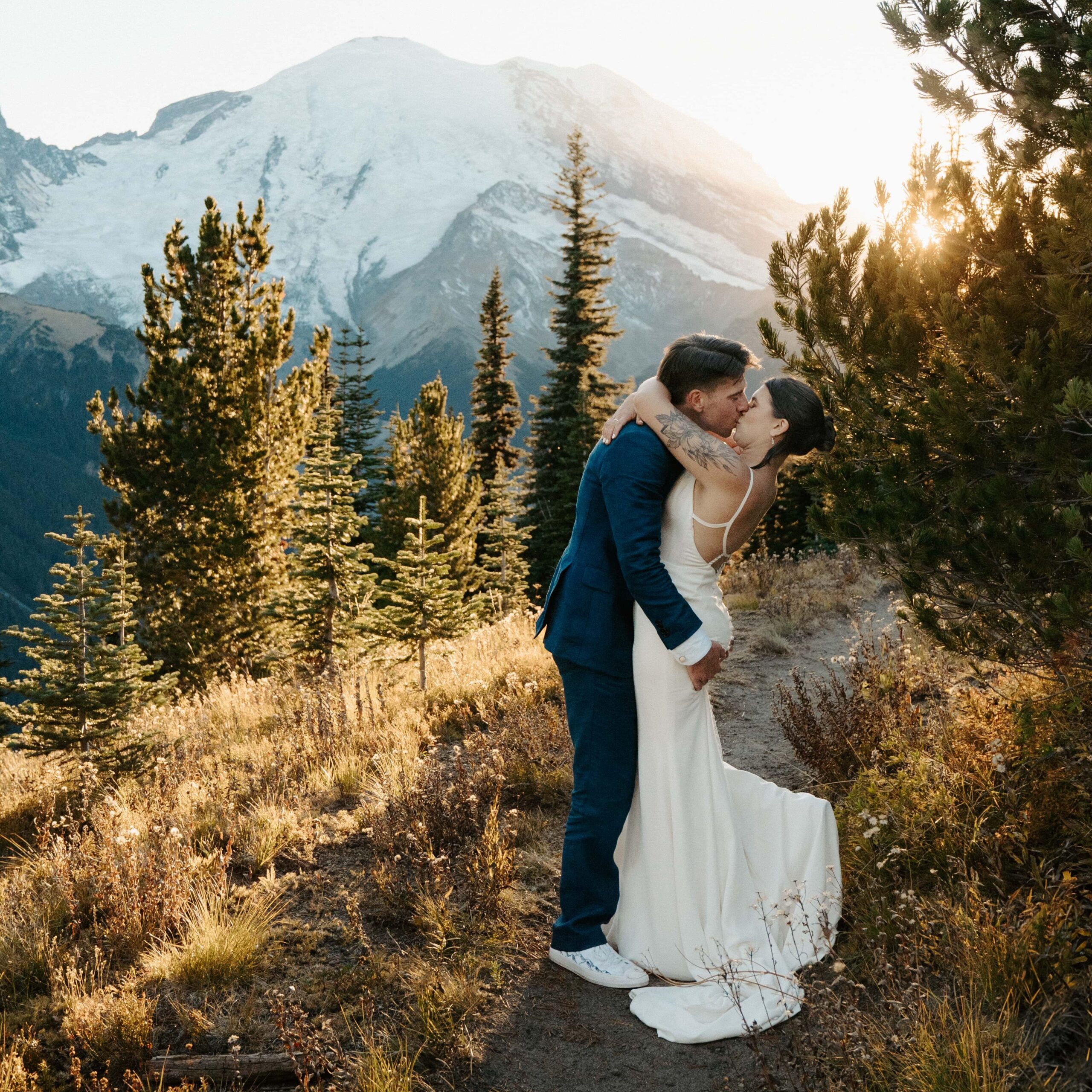 Bride and groom in wedding attire kissing on a trail with the sun setting and Mount Rainier in the background.
