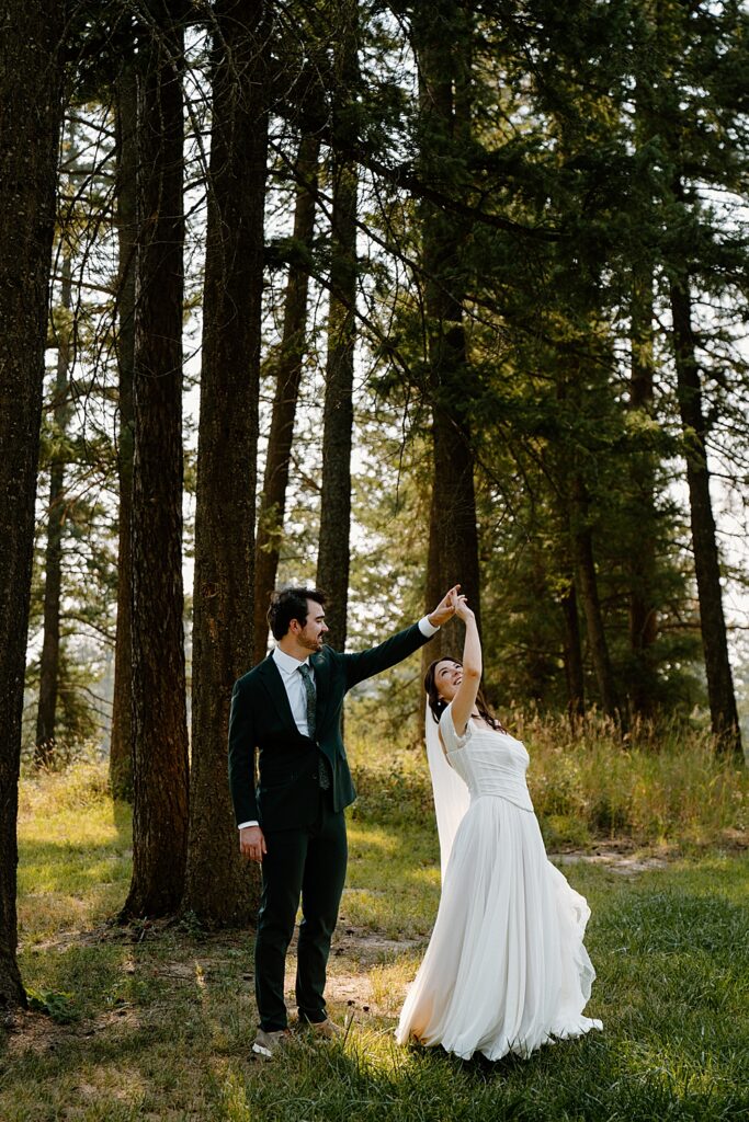 Groom in a green wedding suit twirling bride in a white dress and veil under tall trees in Columbia Falls, MT. 