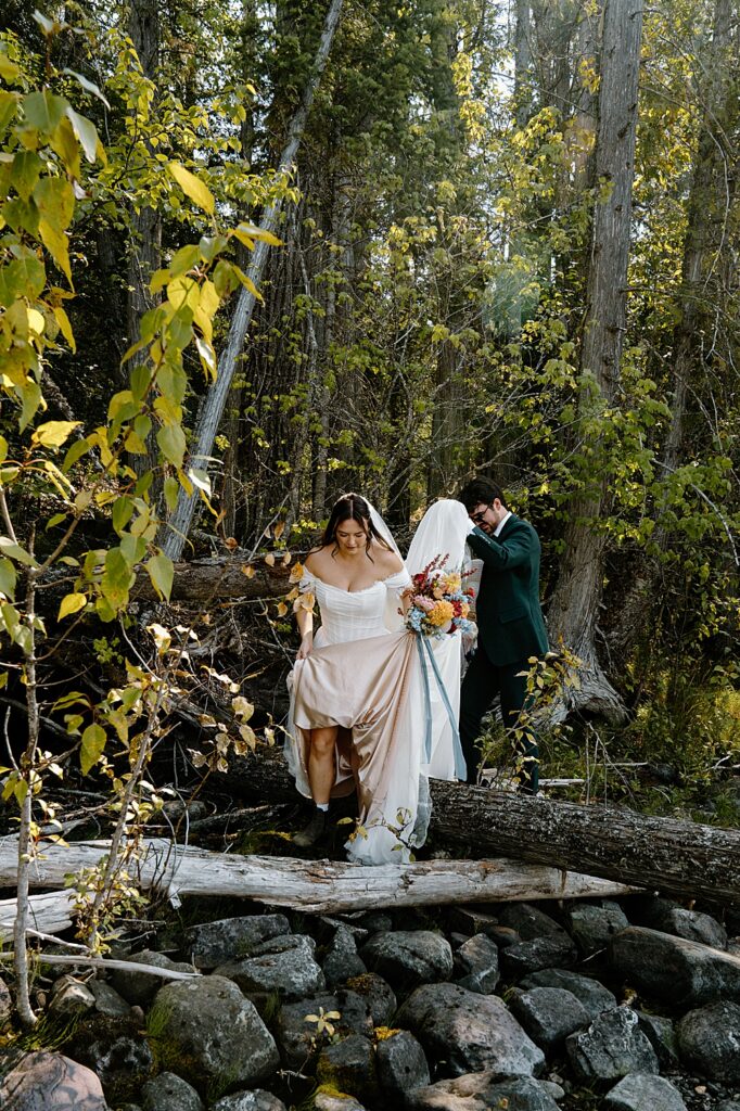 Groom holding bride's wedding train while she holds her bridal bouquet and steps over a log on the rocky shore of Lake McDonald.