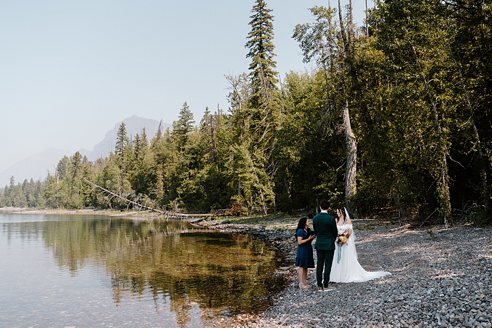 Bride and groom standing on the rocky shore of Lake McDonald having an officiated elopement ceremony at 10-Mile Pullout. 