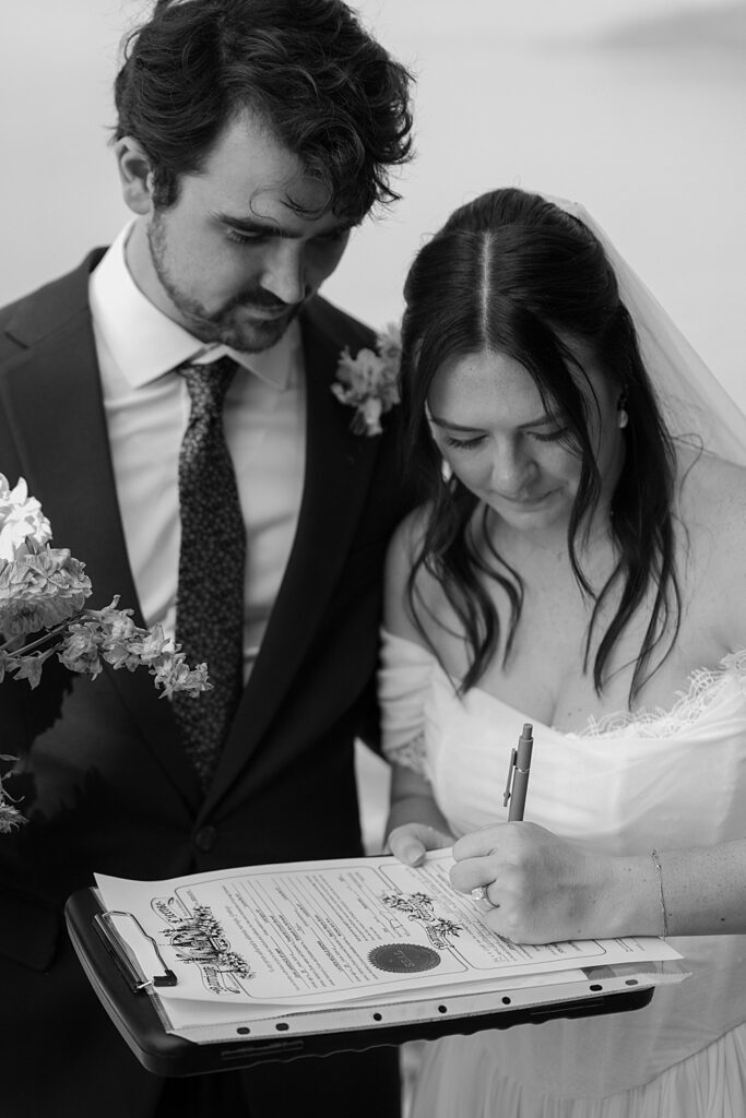 Black & white of a bride signing their Flathead County Marriage License and the groom standing next to her. 