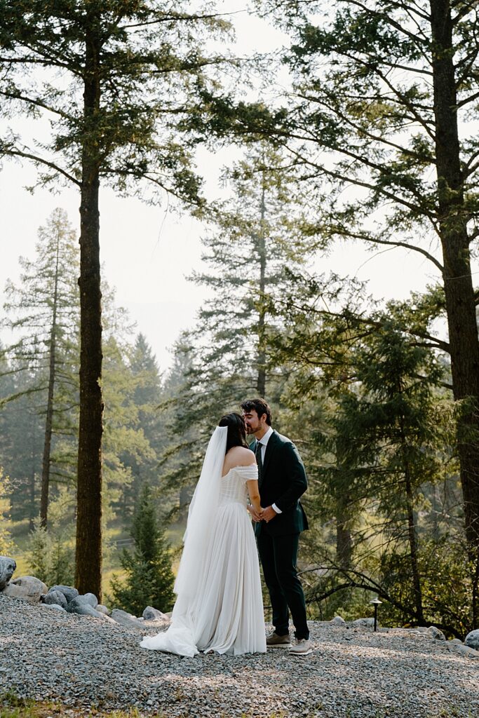 Bride and groom kissing on a gravel path with tall trees behind them. 