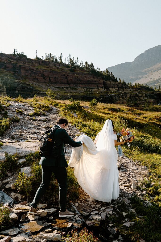 Hiking elopement with groom holding the bride's train on a trail in Glacier National Park. 