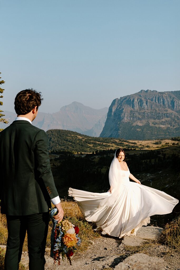 Groom in a green suit holding a colorful summer bridal bouquet facing his bride while she swooshes the train of her wedding dress. 