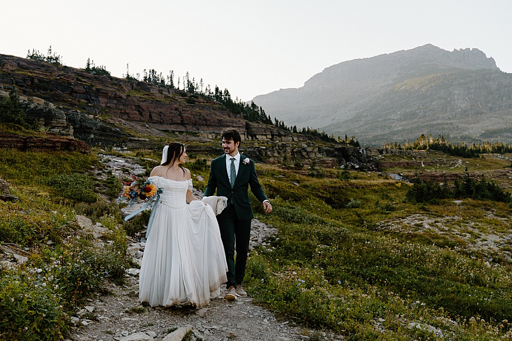 Groom holding bride's train while they hike on a trail at Glacier for their Montana elopement. 