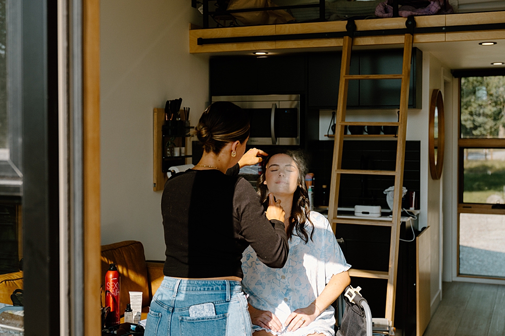 Bride sitting in a chair in a little cabin getting her face sprayed with makeup by an artist from Covet Beauty in Kalispell, MT. 