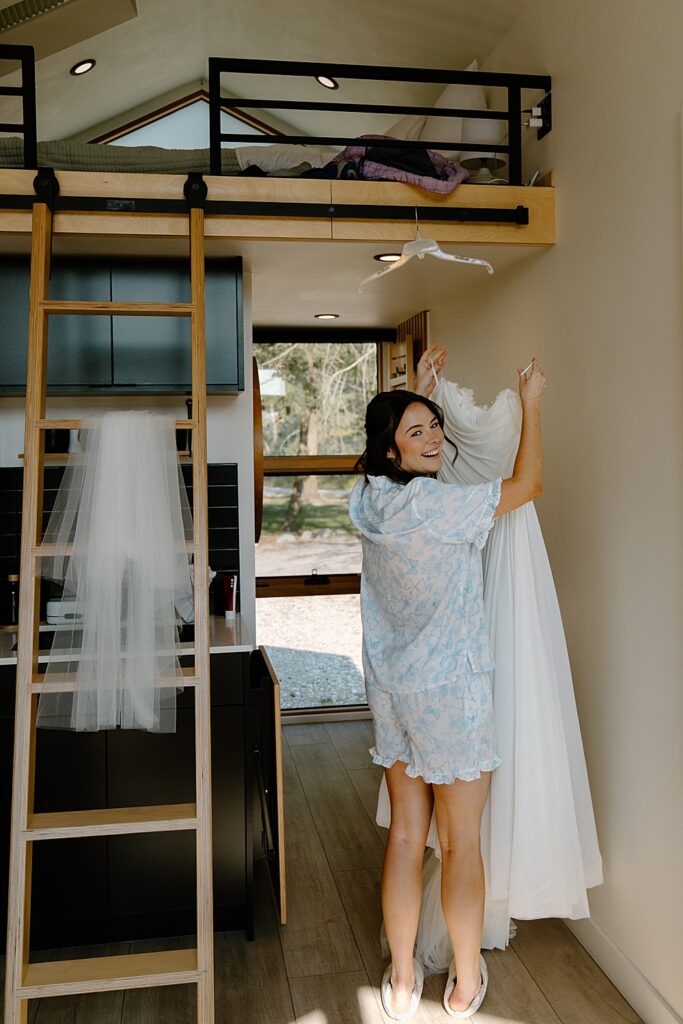 Bride in white shorts and a button-up sleep shirt with blue details, grabbing her wedding dress off the hanger in a cute little tiny home. 