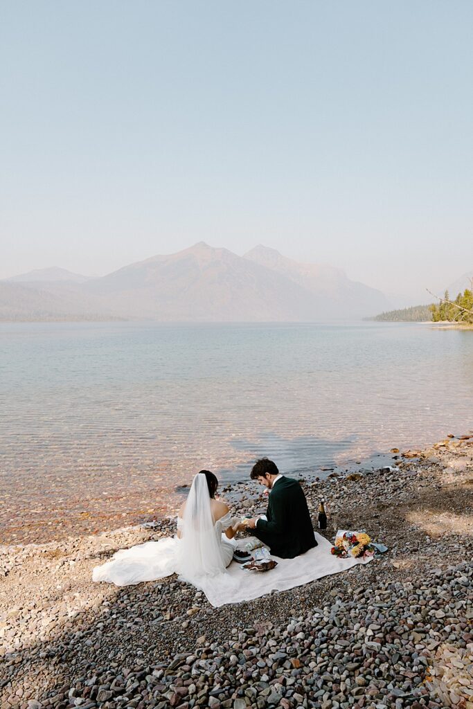 Bride and groom sitting on a picnic blanket on the rocky shore of Lake McDonald for their Glacier Park elopement. 