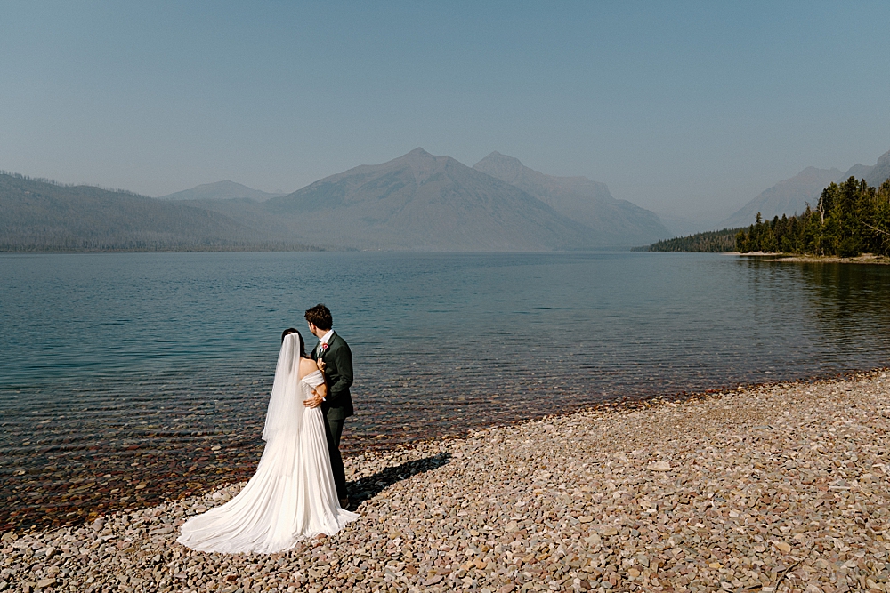 Bride with a white wedding dress and veil and groom in a green wedding suit standing on the beach looking out at Lake McDonald. 