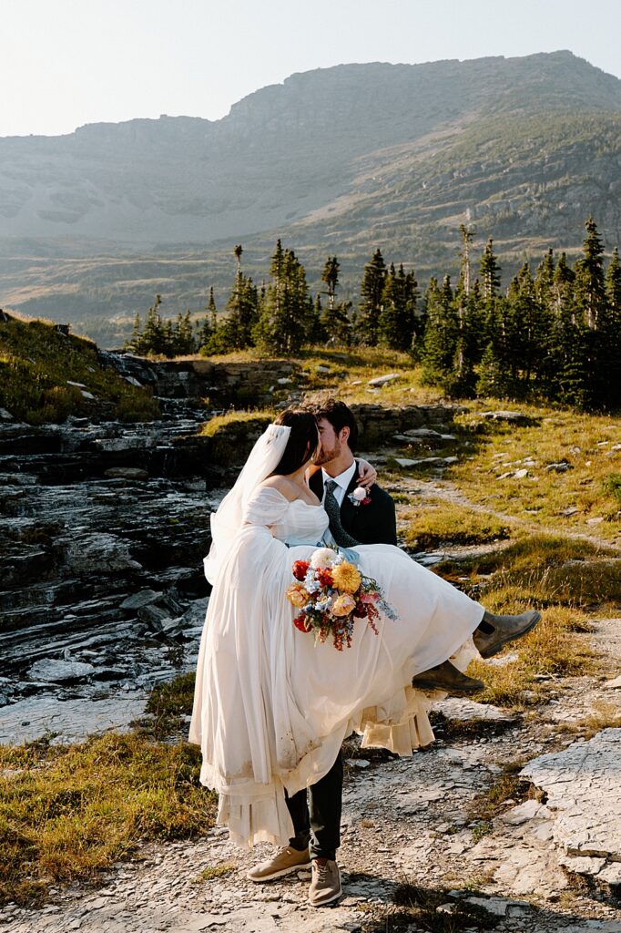 Groom holding bride with colorful wedding bouquet and hiking boots on a trail in Glacier Park.