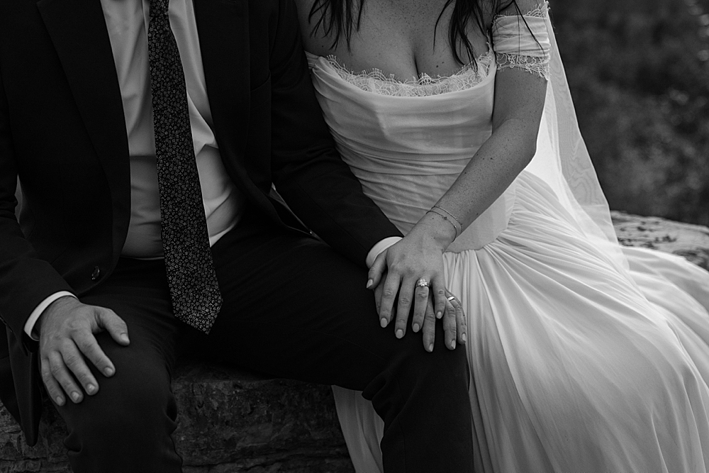 Black and white of bride and groom sitting on a rock with their hands overlapping