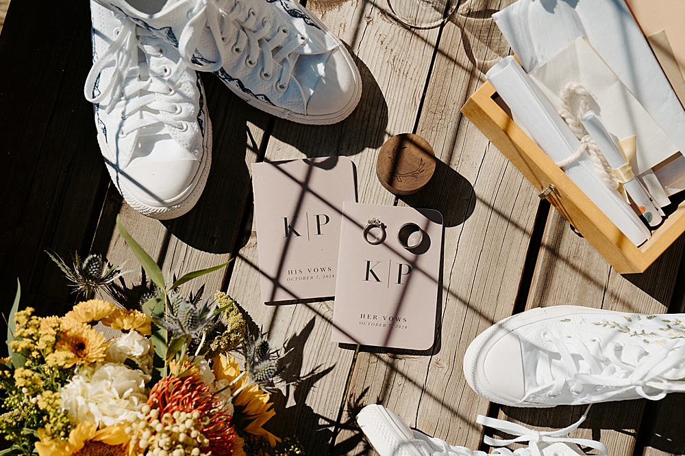 Wedding details set out on a deck with custom vow books, wedding rings, a wooden box filled with letters from loved ones, floral bouquet, and white converse.