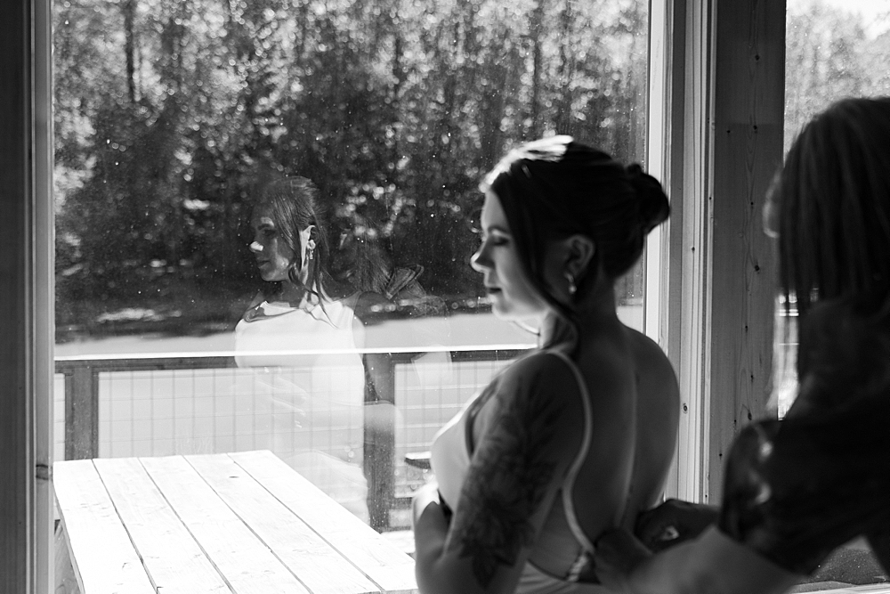 Up close black & white of bride in front of a large window while the mother of the bride is buttoning her dress.